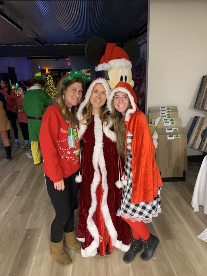 Three women dressed in Christmas costumes posing with a Mickey Mouse caroler decoration.