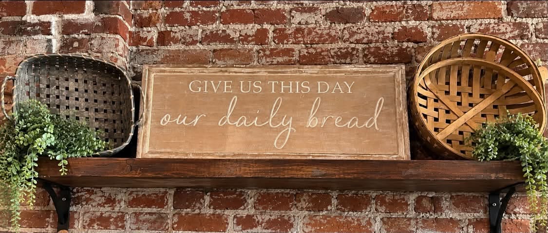 "Give Us This Day Our Daily Bread" wood wall sign on a shelf.